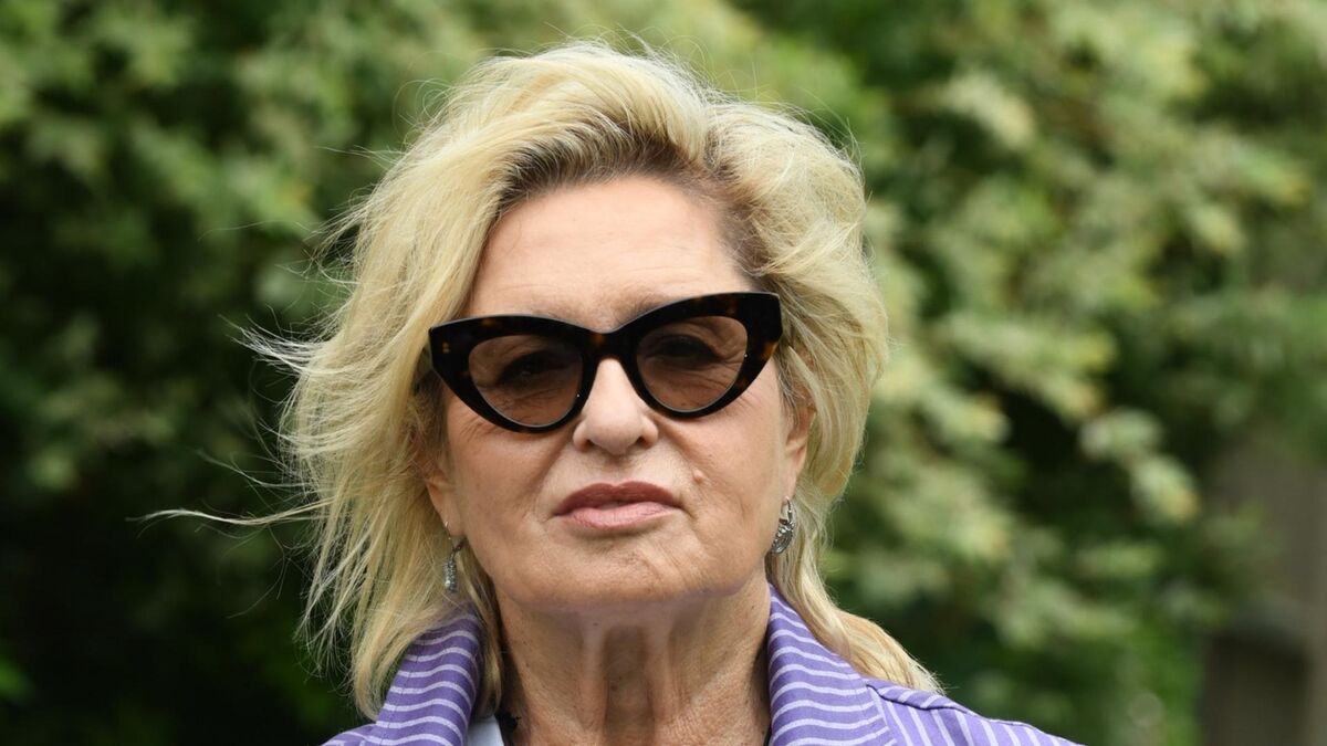 Ewa Kasprzyk patrzy w stronę aparatu, mając na sobie ciemne okulary przeciwsłoneczne i fioletową marynarkę w paski. Ma jasne, krótkie włosy oraz kolczyki. W tle znajduje się rozmyta zieleń drzew i krzewów.
