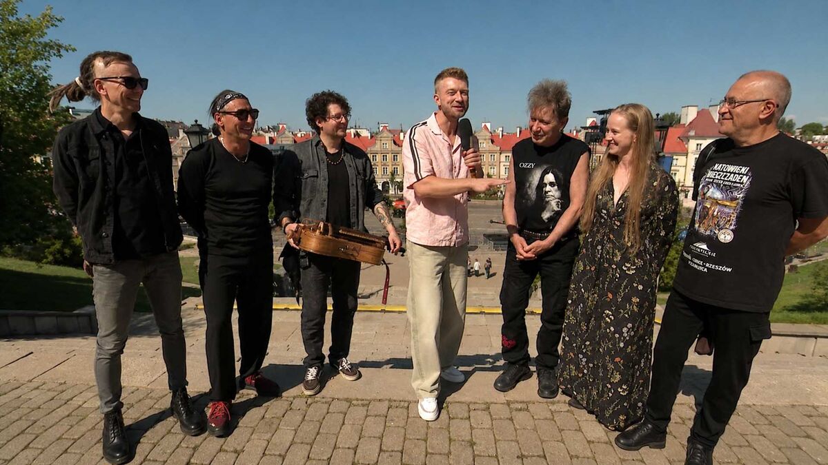 Członkowie zespołu KSU oraz prowadzący stoją na tarasie widokowym podczas plenerowego wywiadu. Jeden z muzyków trzyma instrument strunowy, a prowadzący mówi do mikrofonu, zwracając się do pozostałych uczestników. W tle widać zabytkowe kamienice, brukowany plac i bezchmurne niebo.