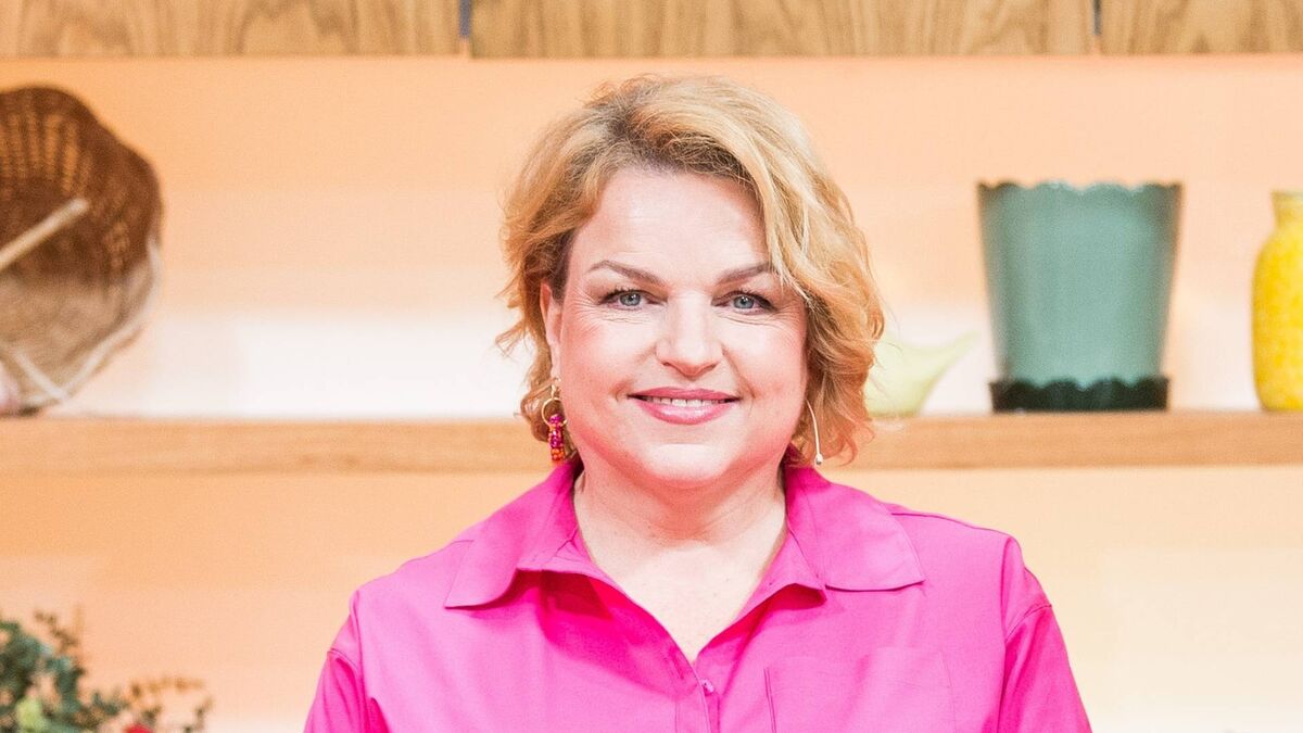 Katarzyna Bosacka uśmiecha się do obiektywu podczas nagrania w studiu. Dziennikarka ma na sobie intensywnie różową koszulę i kolorowe kolczyki, a w tle widać drewniane półki z dekoracjami i doniczkami, tworzące ciepłą, domową scenografię.
