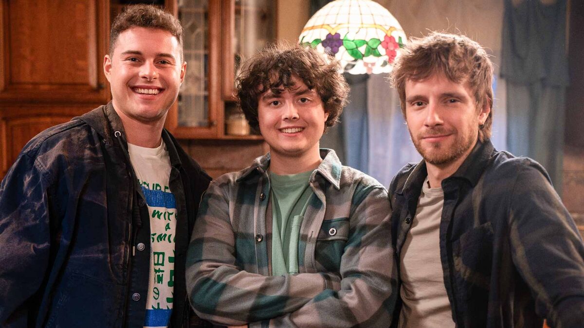 Adam Zdrójkowski, Mateusz Pawłowski, Maciej Musiał, stoją obok siebie we wnętrzu mieszkania z drewnianą zabudową i lampą witrażową w tle. Postacie ubrane są w casualowe stylizacje i ustawione blisko siebie przed kamerą.