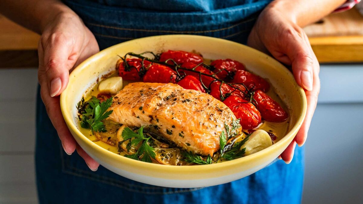 W naczyniu do zapiekania znajduje się filet ryby ułożony na pieczonych warzywach, takich jak marchew, papryka, cukinia i kolba kukurydzy. Danie posypane jest świeżą zieleniną i podane w prostokątnej formie na jasnym blacie kuchennym.