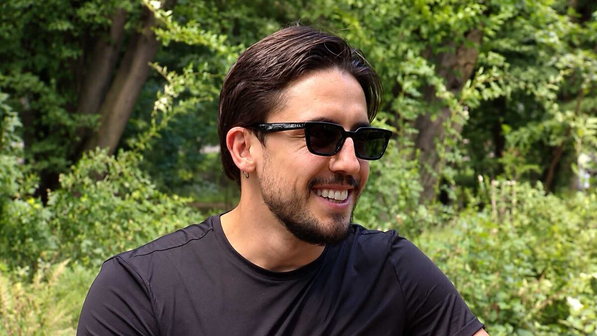 Dawid Kwiatkowski ma na sobie czarną koszulkę i ciemne okulary przeciwsłoneczne. Artysta ma krótki zarost, ciemne włosy zaczesane na bok oraz niewielki kolczyk w uchu. W tle widoczna jest gęsta zielona roślinność i drzewa.