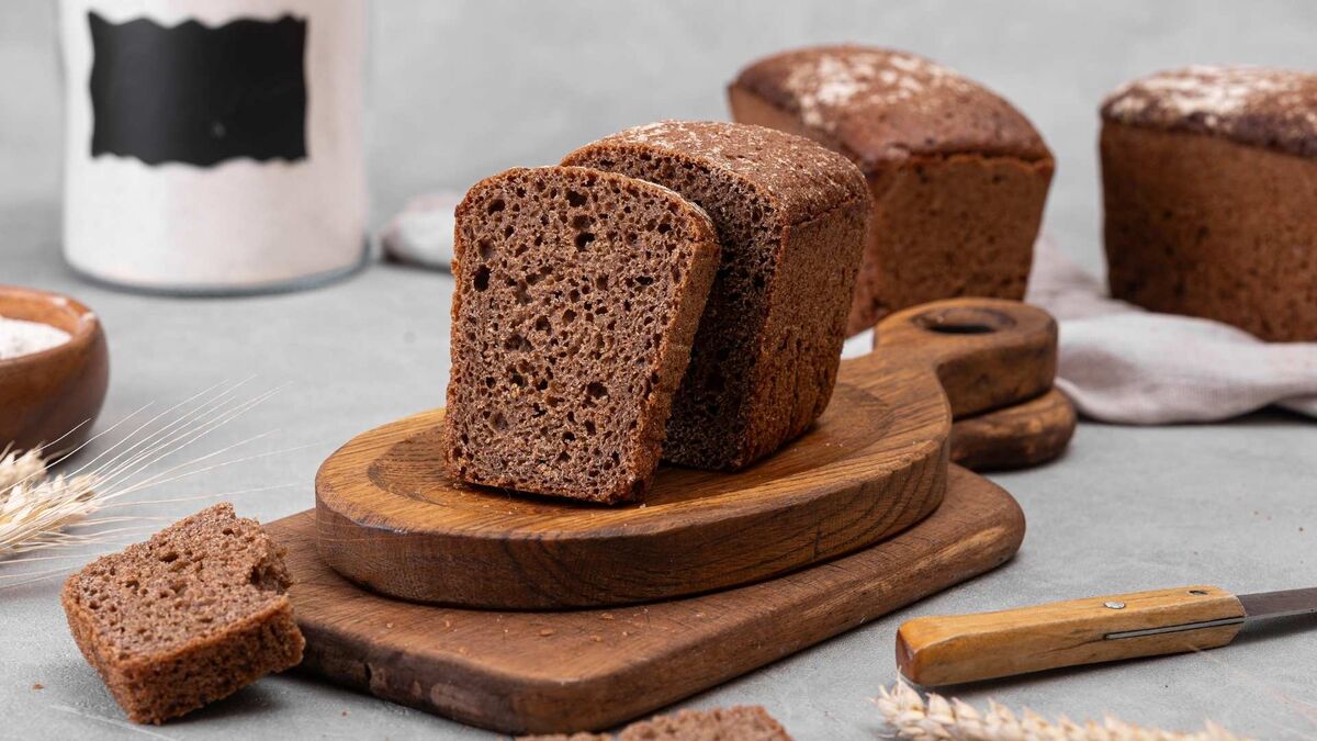 Pokrojony bochenek ciemnego chleba leży na drewnianej desce, obok okruchów i kromki. W tle widać dwa całe bochenki posypane mąką, lnianą ściereczkę oraz szklany słoik z mąką. Na blacie stoją też miseczka z mąką i nóż z drewnianą rączką.