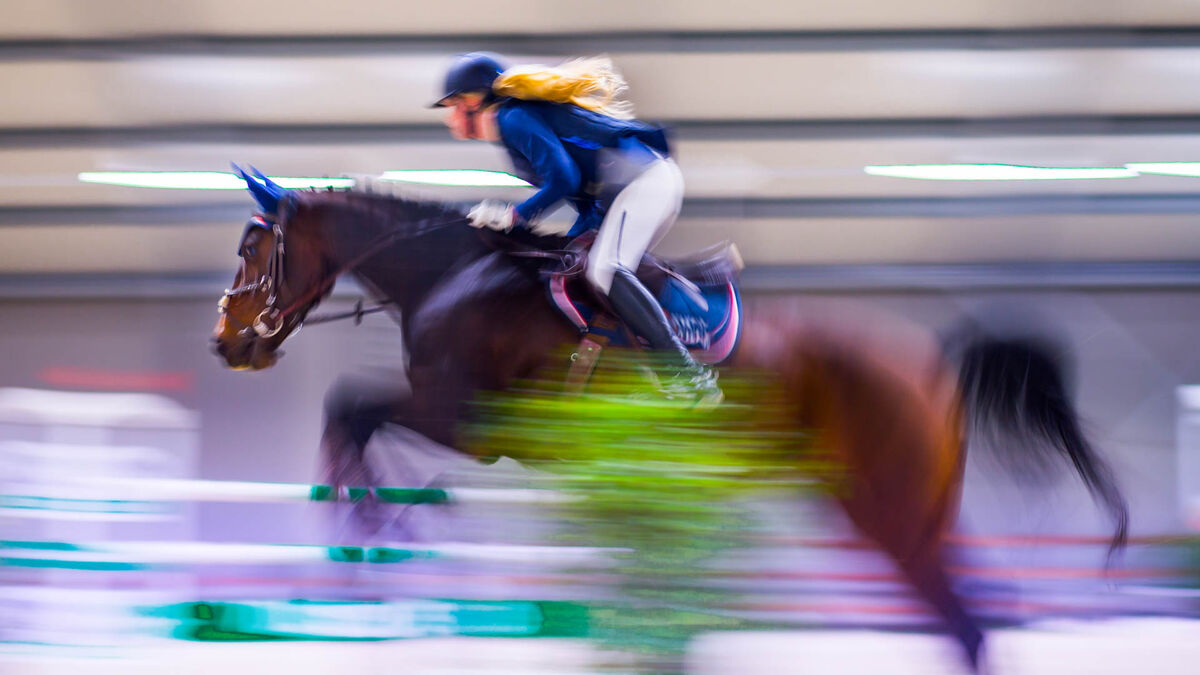 Perlage CAVALIADA Tour. Zawodniczka w kasku i stroju jeździeckim wykonuje skok przez przeszkodę na koniu w hali. Ujęcie z efektem rozmycia ruchu podkreśla dynamikę przejazdu oraz barierki przeszkody na pierwszym planie.