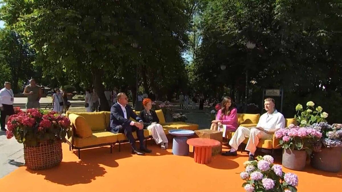 Na zdjęciu widoczne jest plenerowe studio zorganizowane w parku, z żółtymi sofami, stolikami i donicami z kwiatami. Na kanapach siedzą prowadząca oraz troje gości uczestniczących w rozmowie. W tle znajdują się drzewa, alejki parkowe oraz przechodnie.