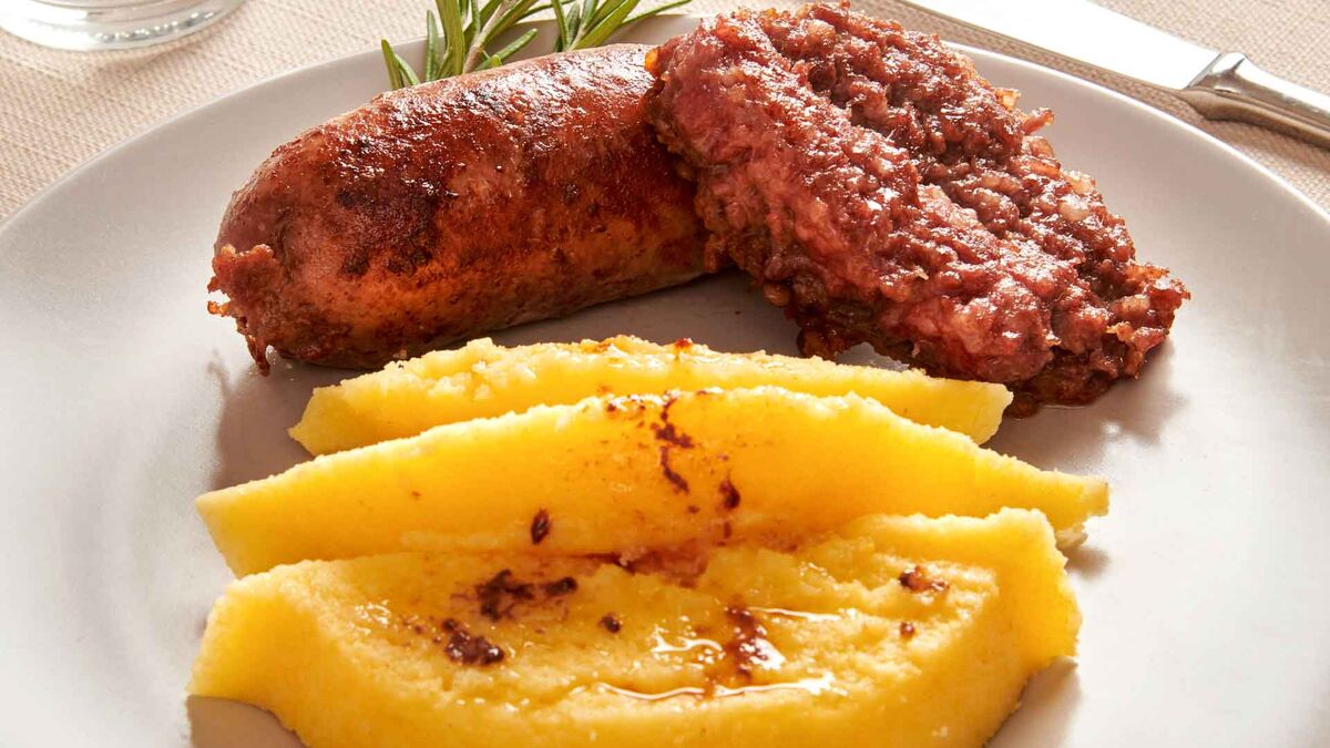 Na białym talerzu podana jest pieczona kiełbasa salsiccia oraz porcja polenty w formie plastrów. W tle widać sztućce i gałązkę rozmarynu, a danie jest ujęte w zbliżeniu.
