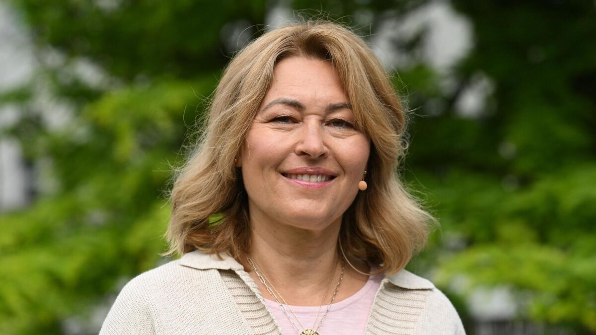 Beata Sadowska uśmiecha się do aparatu, stojąc na tle rozmytej zieleni drzew i krzewów. Ma jasne, sięgające ramion włosy i jest ubrana w jasny kardigan narzucony na różową bluzkę. Na jej policzku widoczny jest mikrofon nagłowny.
