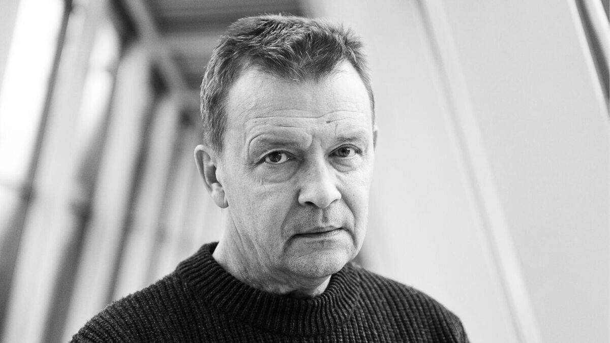 Jan Frycz patrzy wprost w obiektyw z poważnym wyrazem twarzy. Aktor ma krótkie włosy i ciemny sweter z wyraźną prążkowaną fakturą. W tle widoczne są rozmyte jasne ściany i ukośne elementy konstrukcyjne.