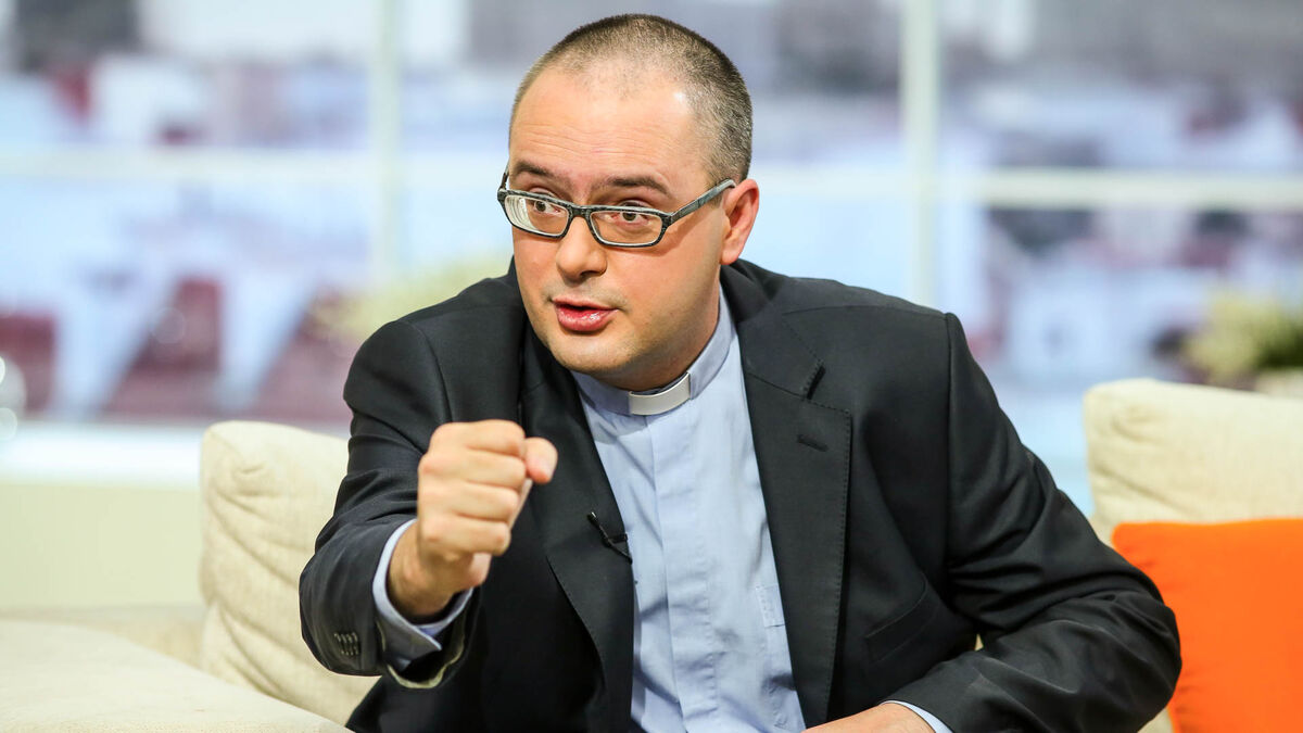Ksiądz Jan Kaczkowski siedzi na jasnej kanapie i gestykuluje prawą dłonią. Ma na sobie czarną marynarkę, jasnoniebieską koszulę z koloratką oraz prostokątne okulary. W tle znajdują się duże okna, jasne elementy wyposażenia i pomarańczowa poduszka.