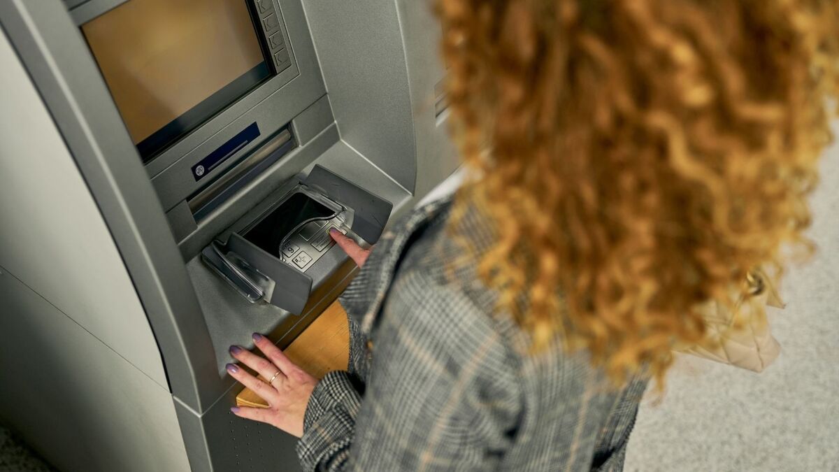 Osoba z kręconymi, rudawymi włosami stoi przy bankomacie i korzysta z klawiatury umieszczonej pod ekranem. Ujęcie wykonane jest z góry i od tyłu, widać szary płaszcz w kratę oraz dłoń opartą o obudowę urządzenia. Po prawej stronie kadru znajduje się jasna torba, a ekran bankomatu pozostaje wygaszony.