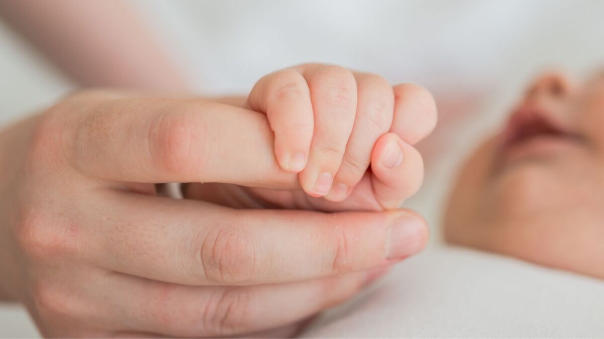 Na zbliżeniu widać dłoń dorosłej osoby, którą chwyta mała dłoń niemowlęcia. Niemowlę ma zaciśnięte palce na palcu dorosłego, a w tle, poza ostrością, widać fragment twarzy dziecka. Zdjęcie utrzymane jest w jasnych, miękkich barwach i pokazuje delikatny kontakt rąk.