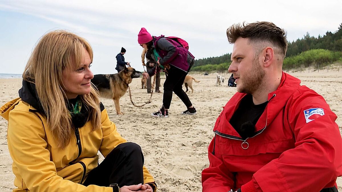Kobieta i mężczyzna siedzą naprzeciwko siebie na plaży i prowadzą rozmowę. Oboje mają na sobie kolorowe kurtki w żółtym i czerwonym kolorze, a w tle widać spacerujące osoby oraz psy. Za nimi rozciąga się piaszczysta plaża i pas zielonych wydm.