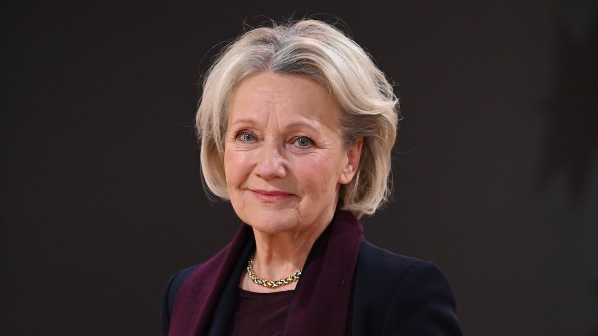 Maria Pakulnis stoi na ciemnym, neutralnym tle z lekkim gradientem po bokach kadru. Ma jasne, krótko ułożone włosy, ciemną marynarka oraz bordowy element garderoby podkreślający linię szyi. Uzupełnieniem stylizacji jest złoty łańcuszek, a kadr pozostaje ciasny i skoncentrowany na twarzy oraz górnej części sylwetki.