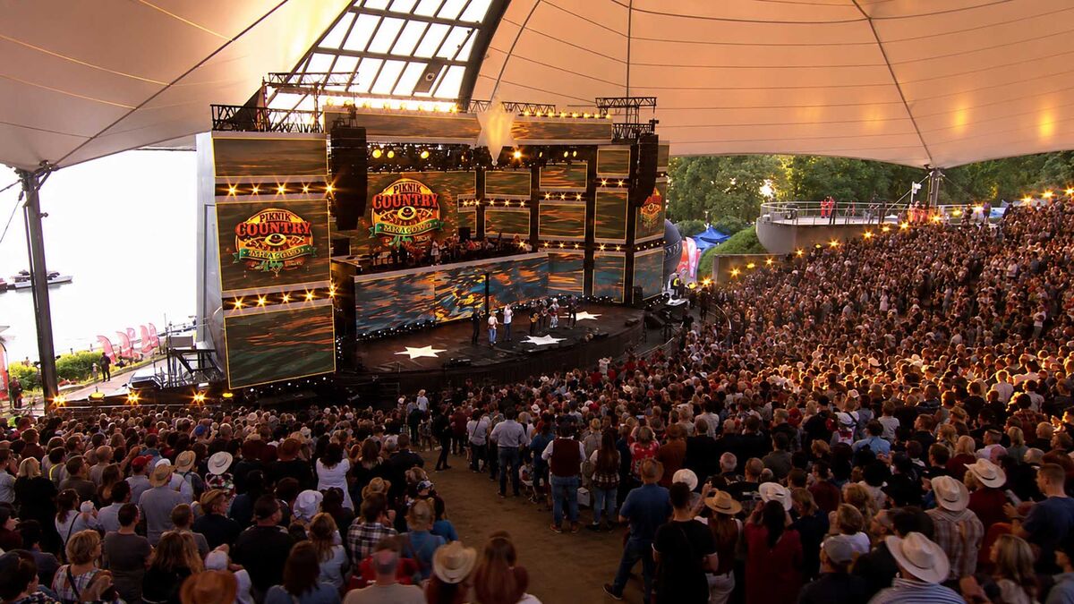 Scena Mrągowo Country and Folk Festiwalu z dużymi ekranami i publicznością zgromadzoną w amfiteatrze. Nad sceną rozpięte jest zadaszenie, a na ekranach widoczne są grafiki wydarzenia. Przed sceną oraz na trybunach znajduje się liczna widownia uczestnicząca w koncercie.