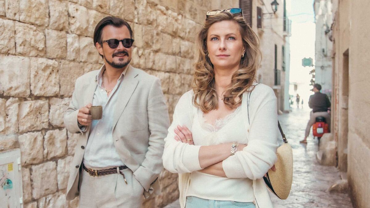 Laura Breszka stoi na wąskiej uliczce obok Dawida Ogrodnika trzymającego filiżankę. Ma na sobie jasną bluzkę i kardigan, a na ramieniu przewieszoną okrągłą torebkę. W tle widoczne są kamienne ściany budynków, brukowana ulica i osoba na skuterze.