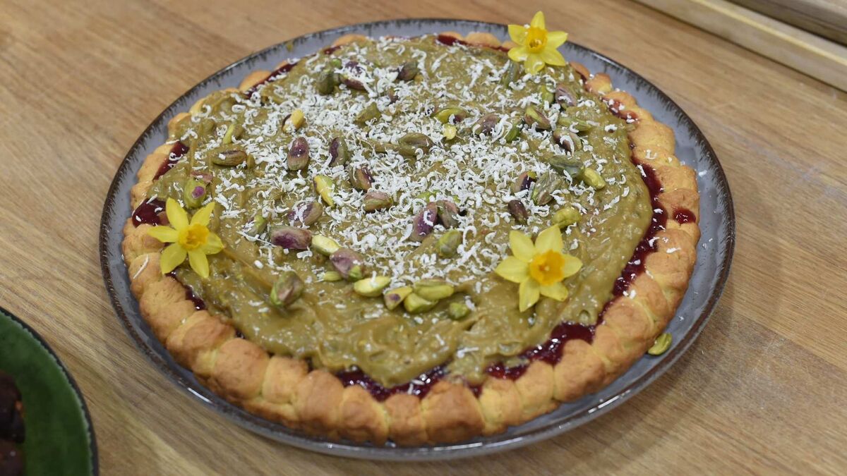 Mazurek pistacjowy na kruchym spodzie. Na wierzchu znajduje się krem pistacjowy, wiórki kokosowe i kawałki pistacji. Ciasto jest udekorowane żółtymi kwiatami.