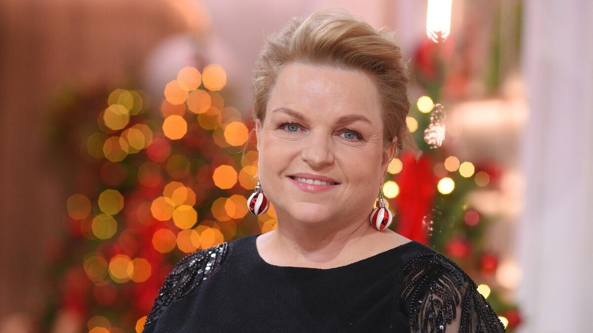 Katarzyna Bosacka uśmiecha się do obiektywu na tle rozmytych, świątecznych lampek i czerwonych dekoracji. Ma krótko upięte blond włosy, czarną sukienkę z połyskującymi zdobieniami na rękawach oraz wiszące kolczyki w biało-czerwone paski. W tle widać miękkie, ciepłe światło i zarys choinkowych ozdób.